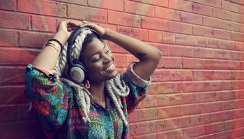 Black woman leaning on brick wall listening to headphones