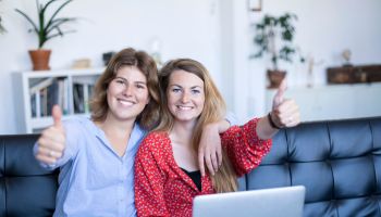 Portrait Of Cheerful Female Friends Gesturing Thumbs Up While Using Laptop On Sofa At Home