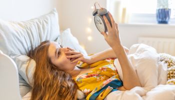 Worried and sick blond young woman in bed late in the morning, overslept looking at the alarm clock ad late for work or school