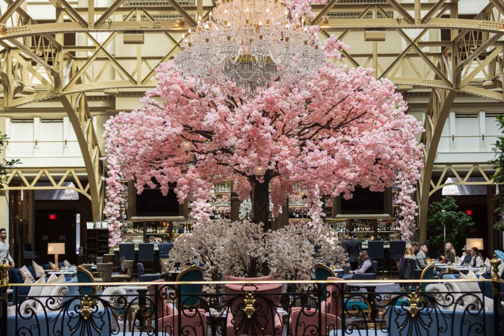A large, ornate chandelier hangs above a lush cherry blossom tree in a grand, open-concept restaurant or lobby. Patrons are seated at tables around the tree.