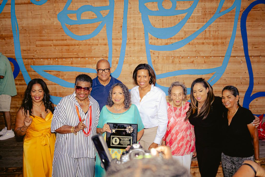 A group of smiling people, including an older woman holding an award, posing in front of a colorful mural.
